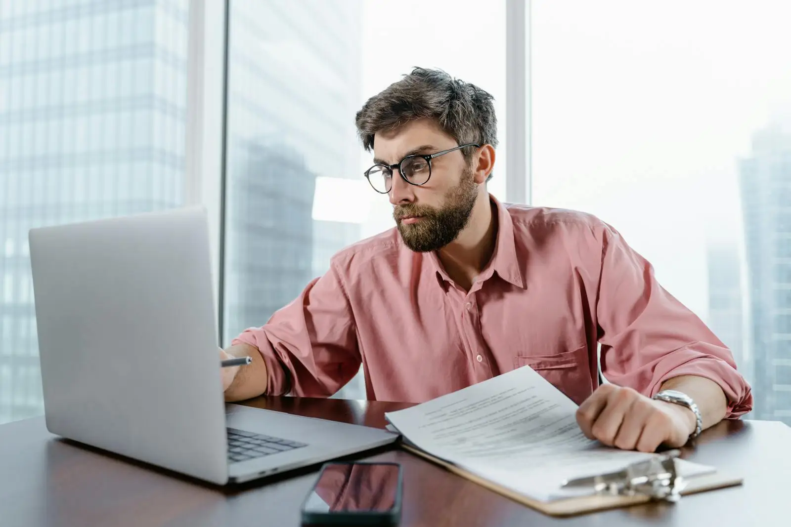 A businessman in an office reviews documents and data on a laptop, concentrating on financial analysis.